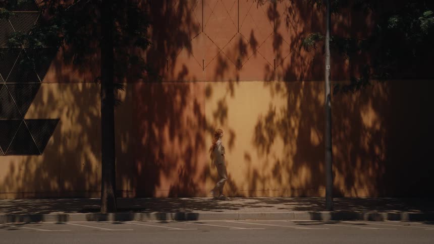 A lone woman walks along a sun dappled sidewalk her figure moving through the shadows cast by trees onto a large textured wall