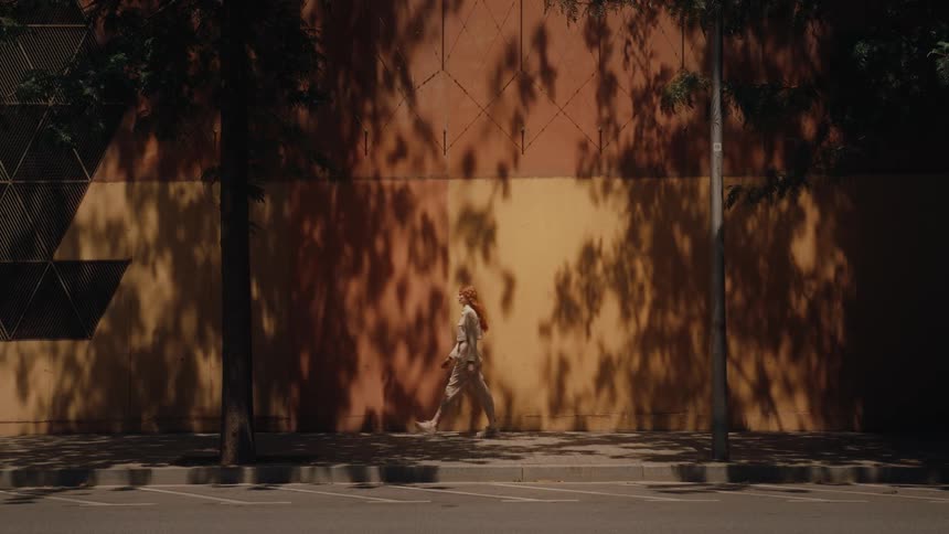 A woman walking on the sidewalk beside a building wall that reflects tree leaves from the sunlight at midday