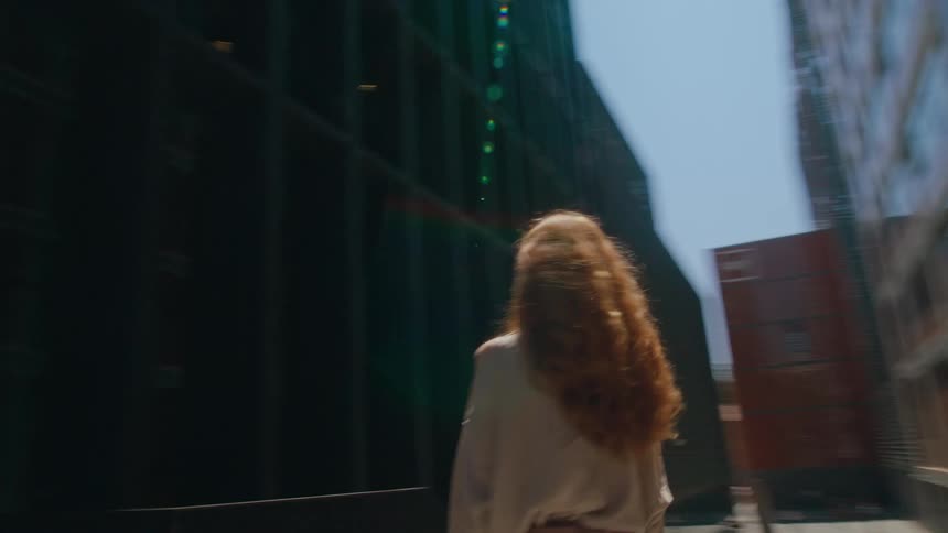 A woman with long flowing red hair and a lightcolored blouse stands outdoors shielding her eyes from intense sunlight while looking upwards