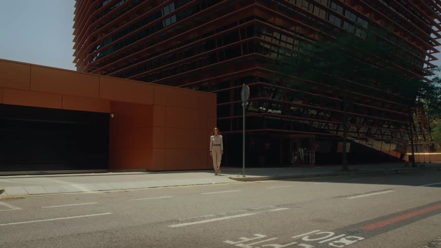 A woman is walking towards the camera on a street next to a modern angular building with orange and glass elements