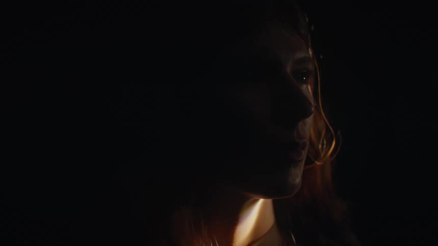 A closeup side profile of a woman with long red hair her face partially obscured by shadow looking to the right as warm light grazes her features