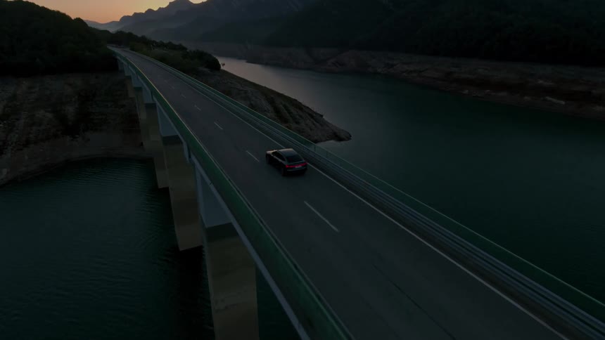 Aerial view of a car driving across a bridge over a reservoir surrounded by mountain hills as the sunsets on the horizon