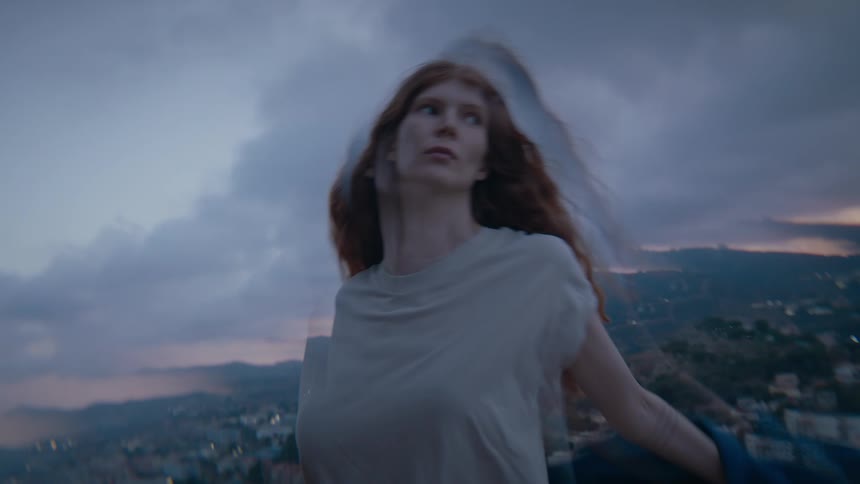 A woman with long red hair and a blue jacket stands in front of a hazy urban landscape under a cloudy sky