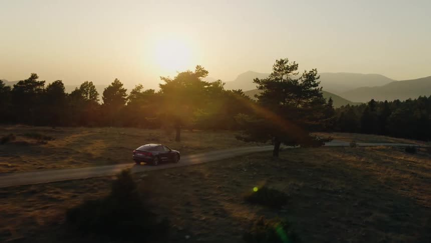 A car drives along a winding rural road through a grassy forest at sunset