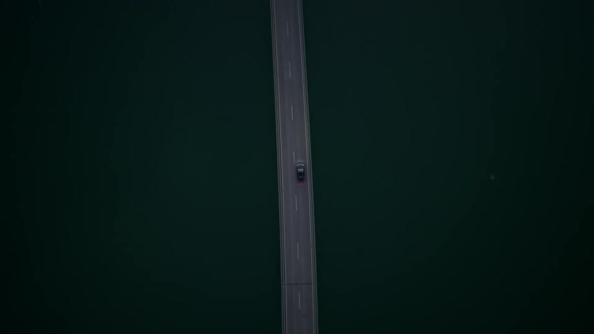 A car crosses a narrow bridge over water and rocks seen from above