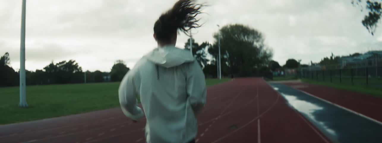 A woman jogs alone on a wet outdoor track under a cloudy sky