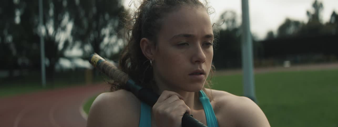 A young lady in activewear holds a pole vault pole on the track field at midday