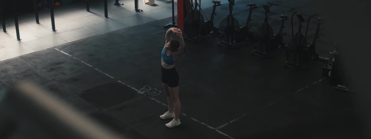A young woman in workout clothes stretches her arms overhead in a gym with dark flooring and various exercise equipment