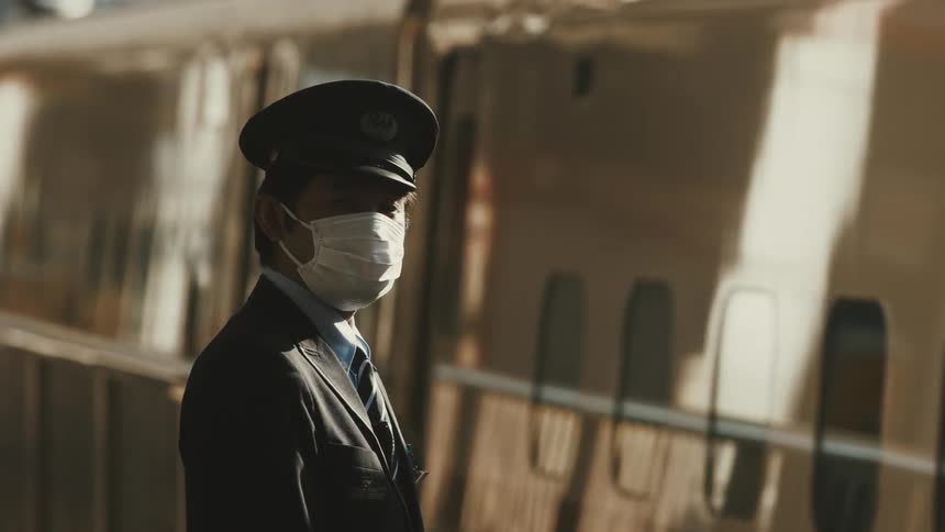 An asian man wearing a military uniform mask and hat stands beside a train coming to a stop at a train station