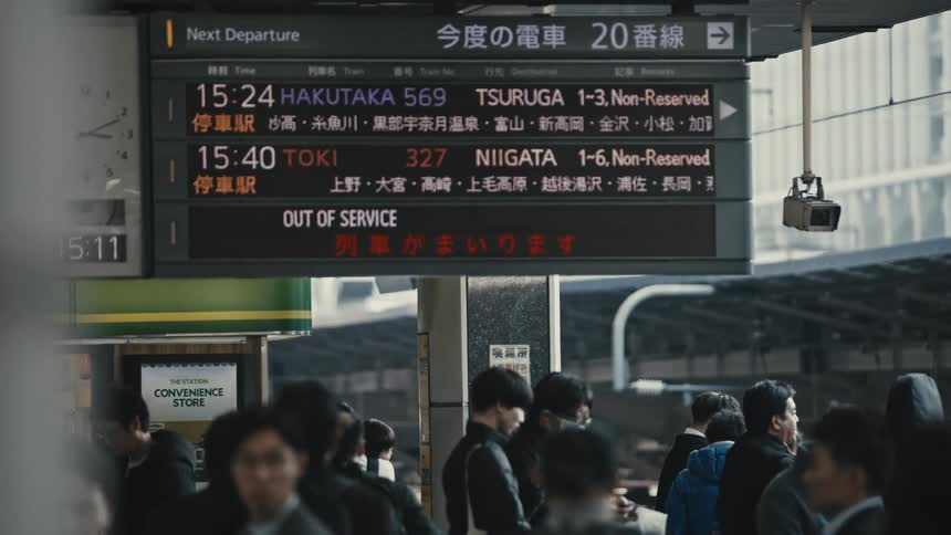 A large group of people stands waiting for the train at a train station with a screen displaying the train schedule