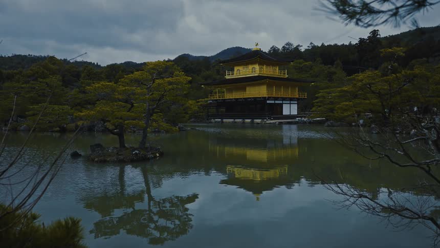 Mostly yellow building with traditional asian architecture stands on a lake among a grove of trees under an overcast sky