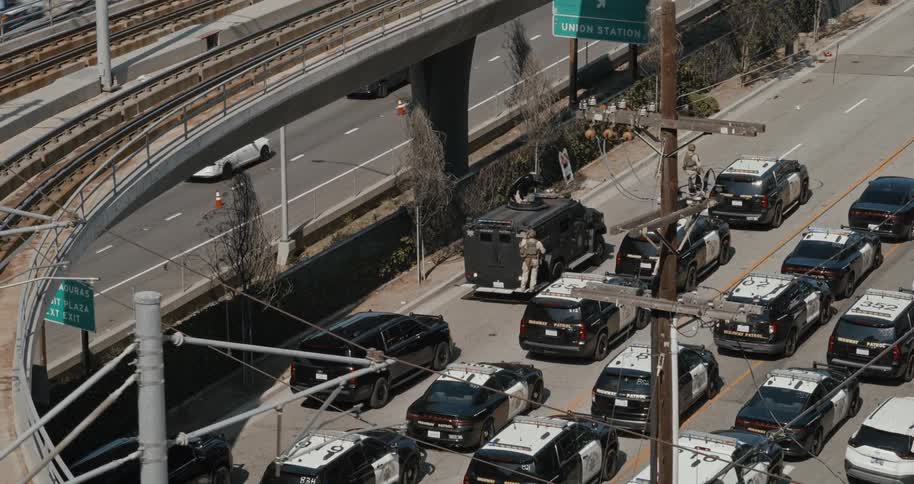 Several police vehicles and officers are gathered on a street near an overpass with some officers escorting an armored vehicle
