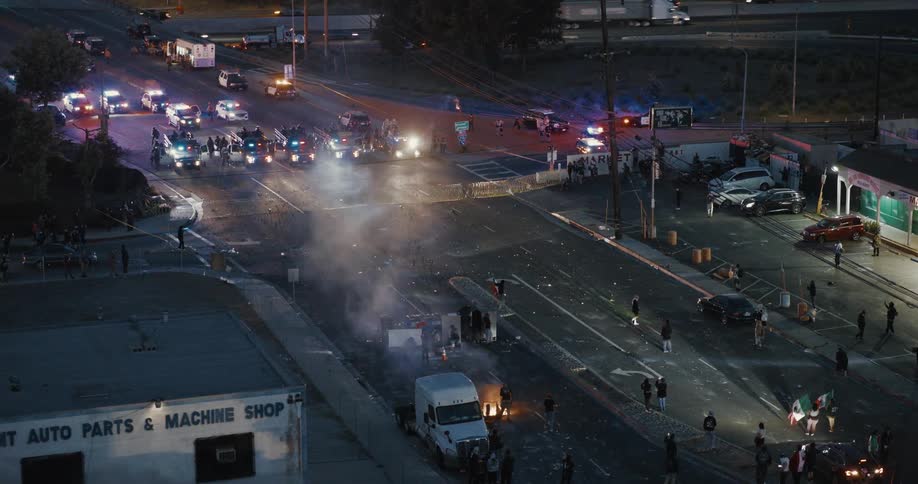 A line of police vehicles and officers blocks off a street at dusk as protesters gather nearby and smoke rises from a fire