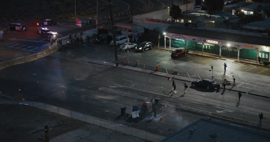 Police respond to a nighttime incident near a parking lot as bystanders and vehicles surround the scene