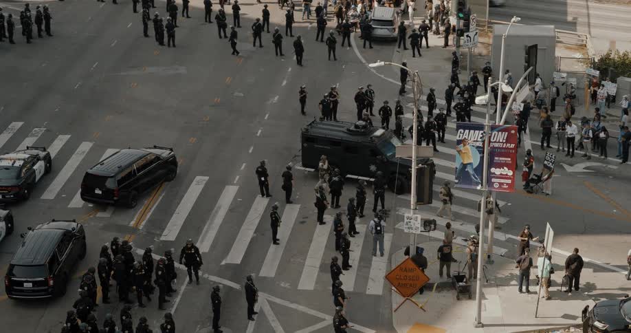 From an aerial perspective dozens of police officers in riot gear assemble at a city intersection with an armored vehicle and patrol cars starting to advance under a highway overpass