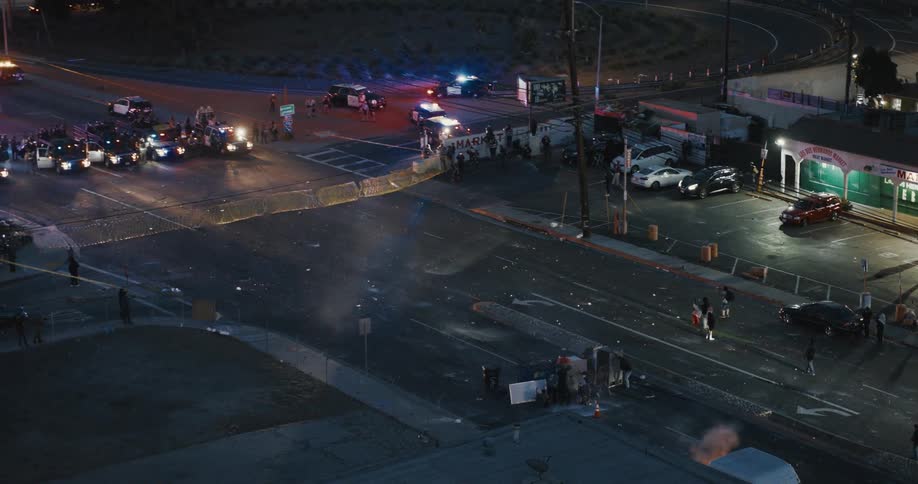 A large number of emergency vehicles including police cars and fire trucks are gathered at an intersection with flashing lights and a crowd of people nearby suggesting a significant incident or response