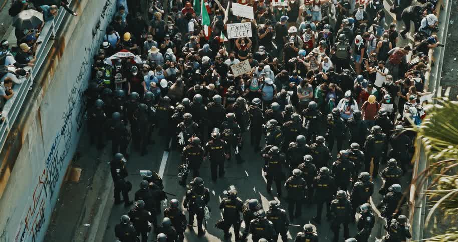 A large group of protesters holding signs and flags confronts a line of riot police on a street next to a graffiticovered wall