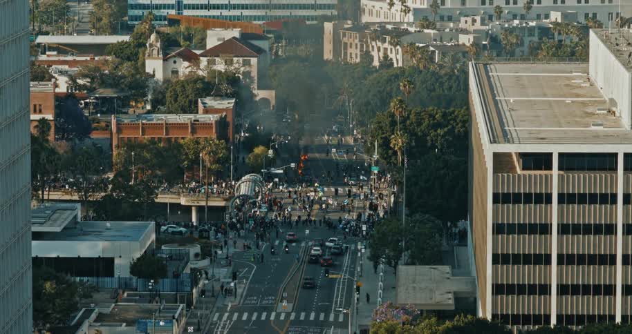 Protestors walk through the streets of a city as Waymo cars burn in the background