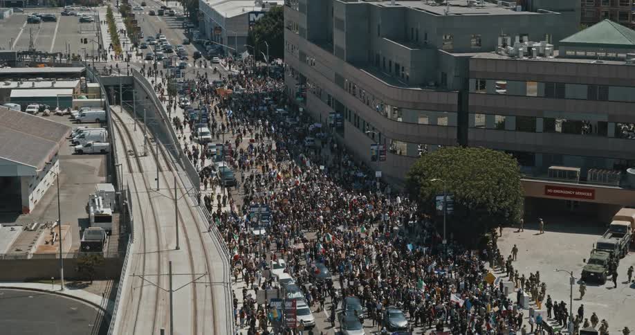 A crowd is forming along a downtown city street with people holding signs and flags beside a building where military personnel are blocking the sidewalk near the entrance