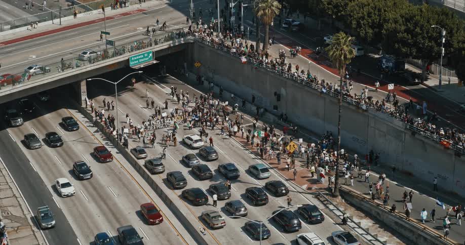 A large group of people are gathered on and around a busy multilane highway causing a significant traffic jam with many vehicles at a standstill