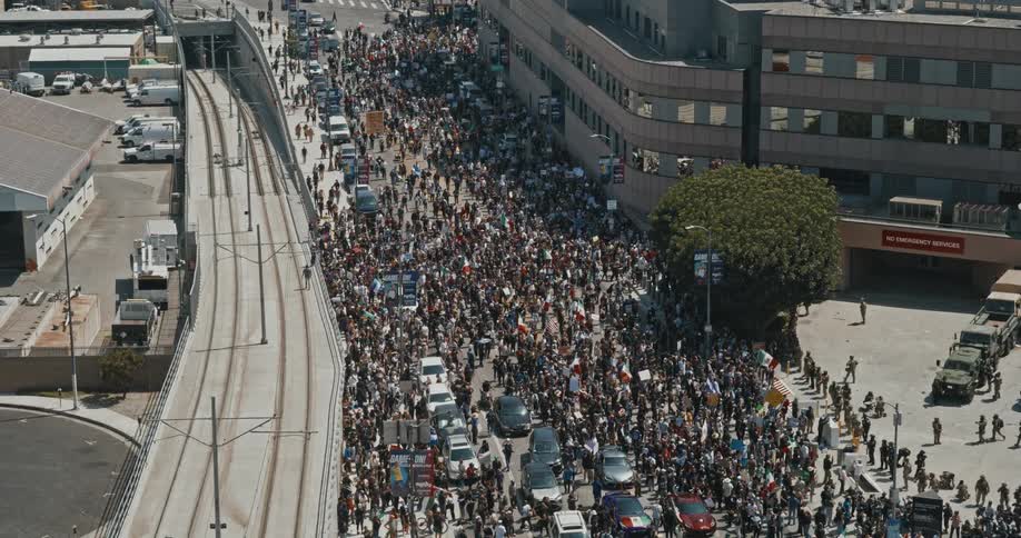 Aerial view of a downtown city street filled with people holding signs and flags beside a building where military personnel are blocking the sidewalk near the entrance