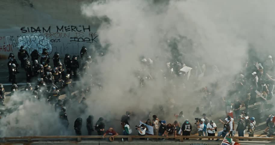 A group of protesters and police officers are clashing on a bridge with smoke filling the air and graffiti visible on the walls