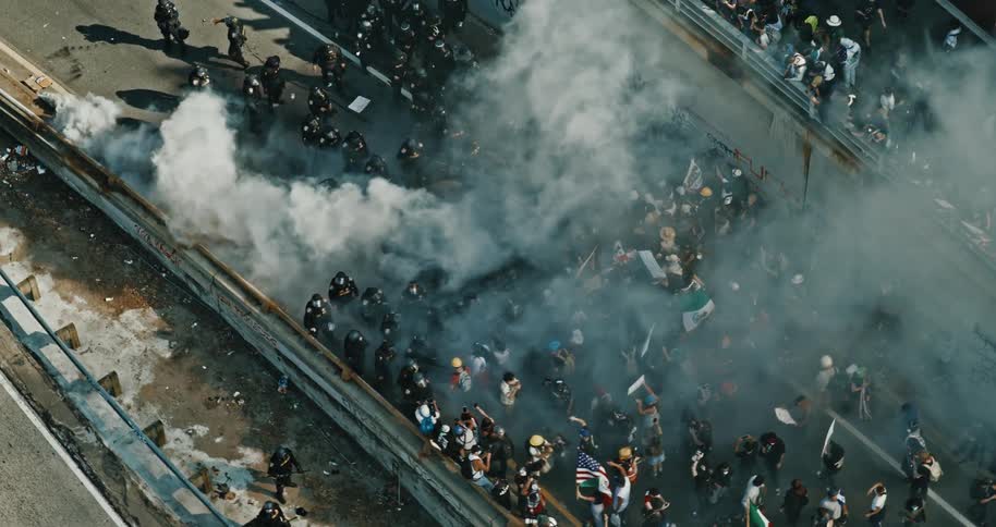 Aerial of a city street filled with protesters and surrounded by law enforcement shrouded in tear gas during daytime