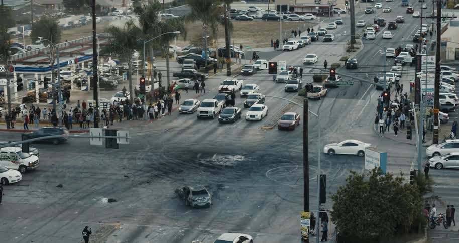 Crowds gather at a chaotic intersection with a burnt car and stalled traffic after a protest