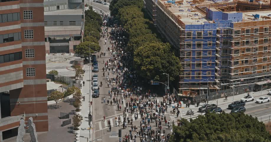 A large crowd of protesters marches down a city street in daylight captured from an aerial view