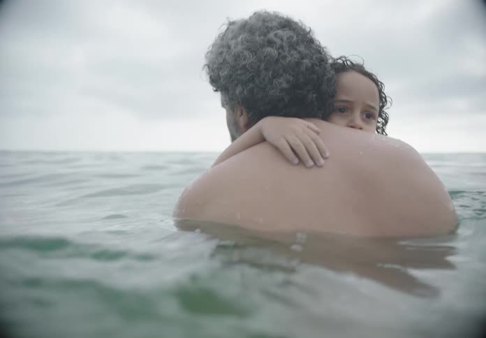 A man with curly grey hair seen from behind embraces a child with dark curly hair in the ocean with the water reaching their chests under an overcast sky