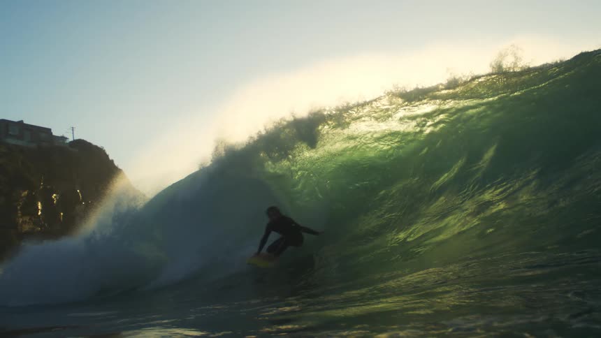 A man surfs a huge ocean wave next to some houses on a hill