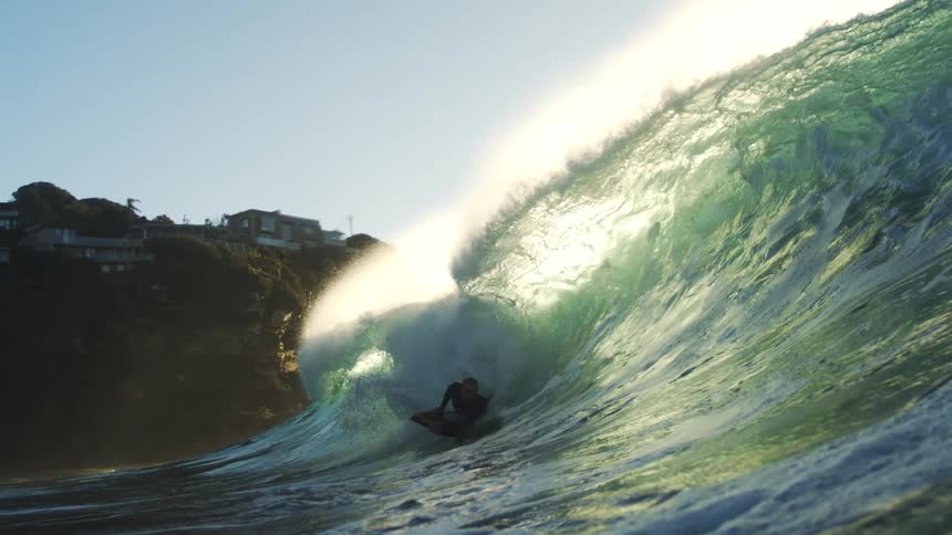 Two men surf a gigantic ocean wave next to a hill with houses on top