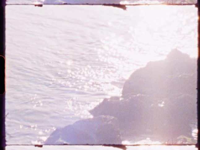 A rocky shoreline is illuminated by bright sunlight with waves gently crashing against the rocks