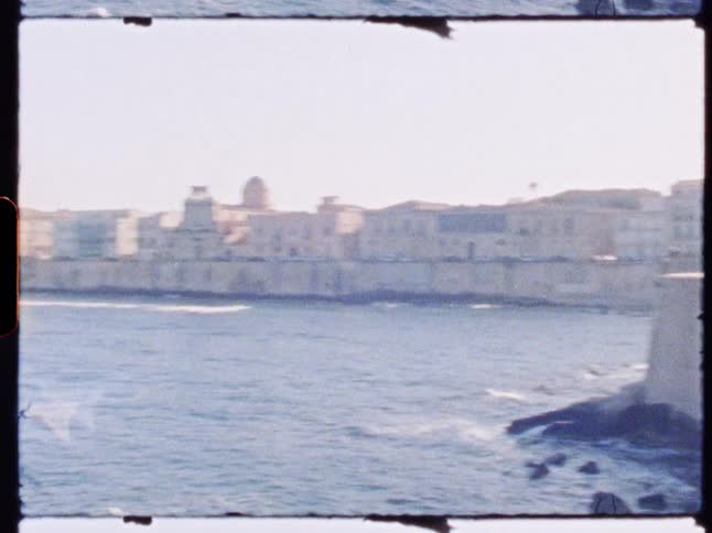 A coastal cityscape features historic buildings and a fortified sea wall overlooking jagged rocks along the shoreline