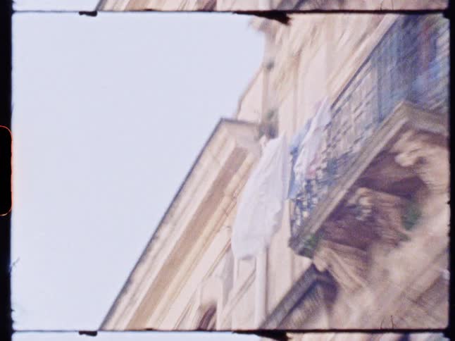 Laundry hangs from an ornate iron balcony on a classical building with intricate architectural details set against a bright clear sky