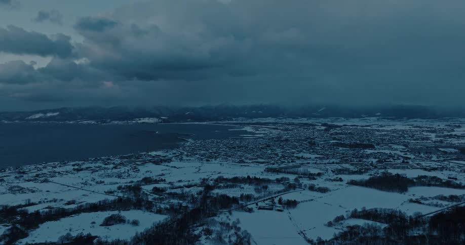 An expansive aerial view captures a snowcovered town nestled between a dark body of water and distant mountains under a cloudy sky at twilight