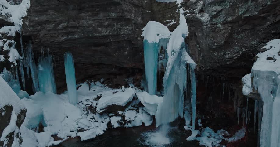 An aerial shot lowers down into a snowy rock cove where a waterfall partially frozen into massive icicles continues to flow into a dark pool below