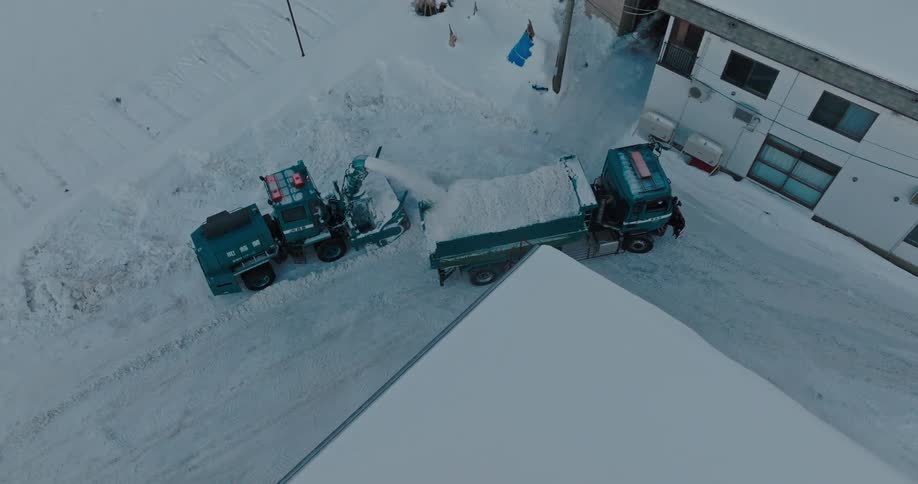 A snow plow blows snow into the back of a dump truck