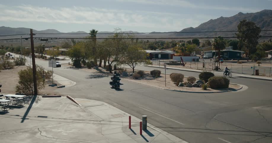 Two people are riding motorcycles on an asphalt street and then turning onto a concrete sidewalk