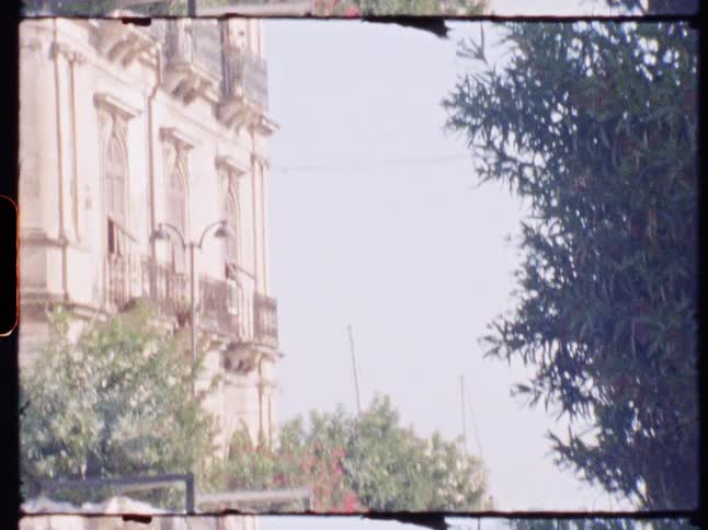Archival footage with a visible film border pans down from an ornate building to reveal a sunny coastal street scene