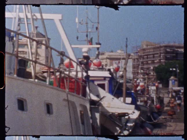 Several large and small boats are docked in a harbor with buildings and a bustling street visible in the background under a clear sky
