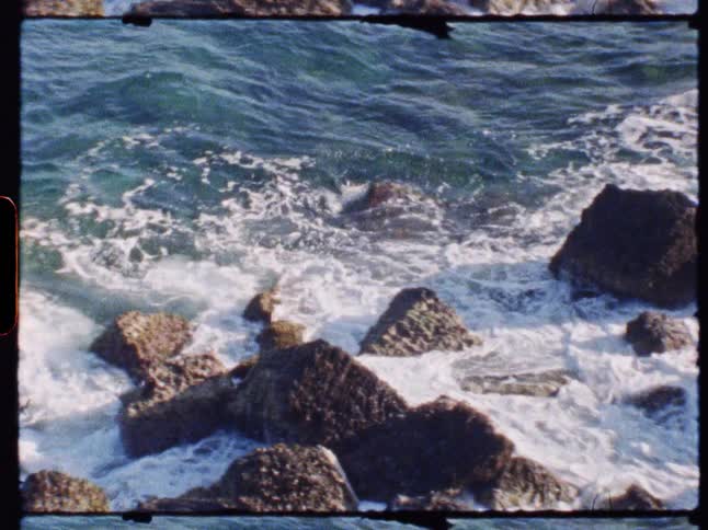 Archival footage with a visible film border shows ocean waves crashing and churning around large rocks on the shore