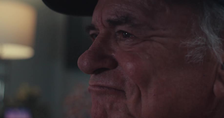 An older man with graying hair and a cap smiling as he briefly removes and then replaces his eyeglasses