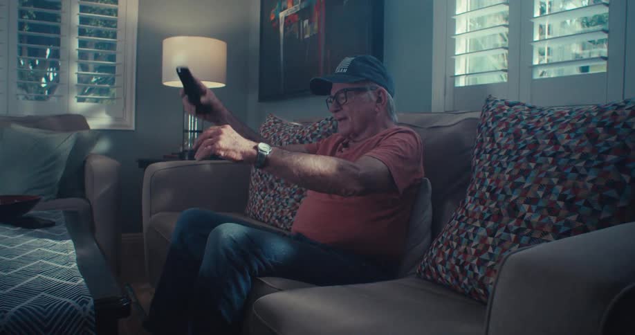 An elderly man in a cap and glasses sits on a couch holding a remote control with his arm extended illuminated by light streaming in from a window with white shutters