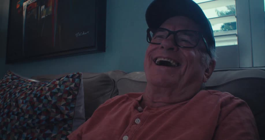 An older man in glasses and a cap seated comfortably on a sofa leans back and bursts into laughter while watching something on tv