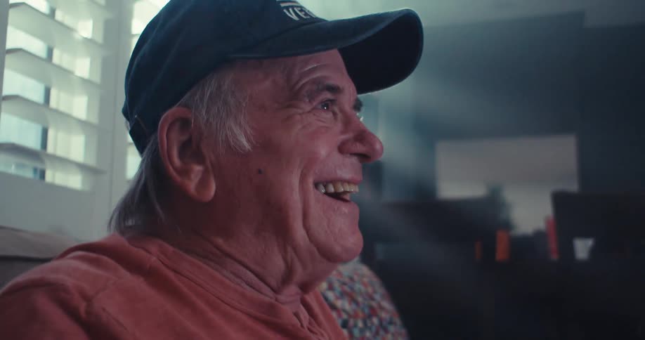 An elderly man wearing a baseball cap and a pink shirt laughs while playing videogames at home illuminated by strong light coming through white window blinds