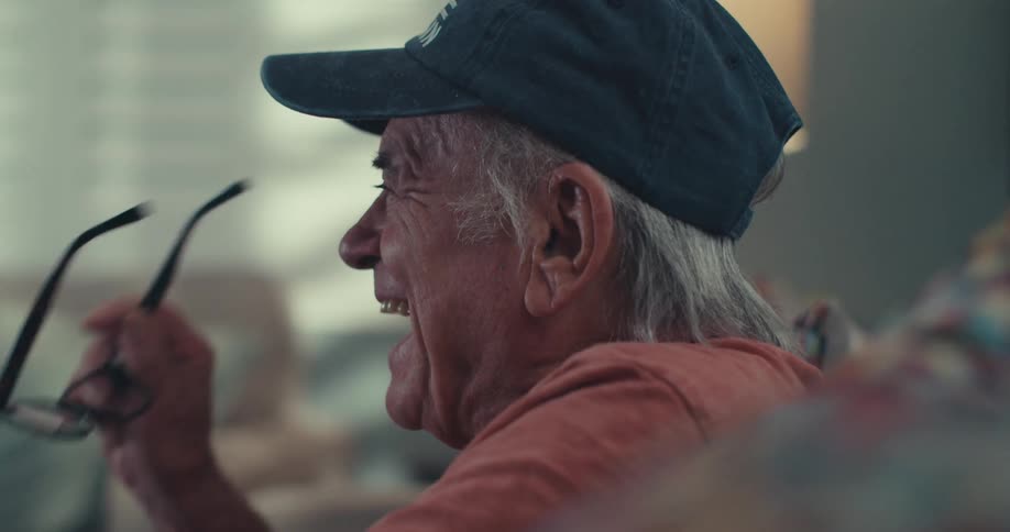 A closeup side profile of an older man in a blue baseball cap captures him laughing with his mouth open while watching television