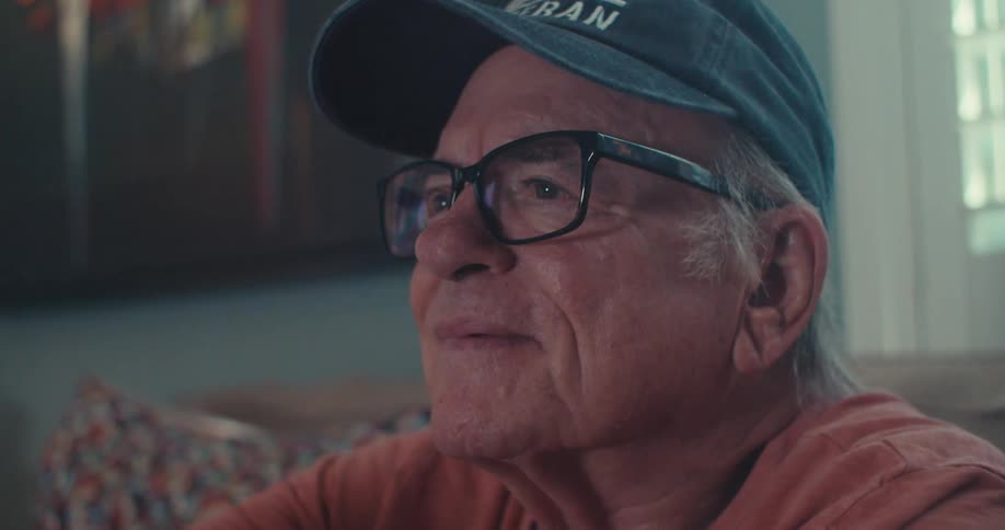 An elderly man wearing a baseball hat and glasses sits on a couch smiling as he watches tv