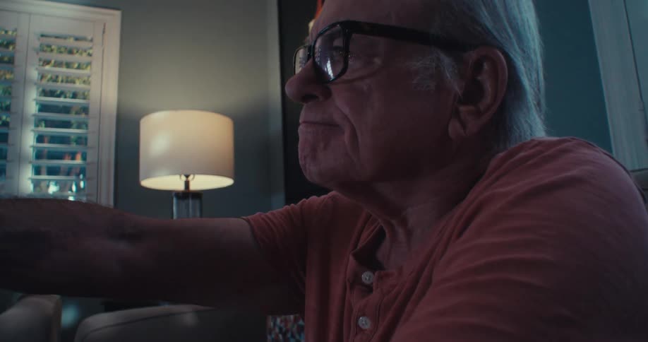 The face of an elderly man who is wearing reading glasses and he is pointing the remote control forward trying to change the channel