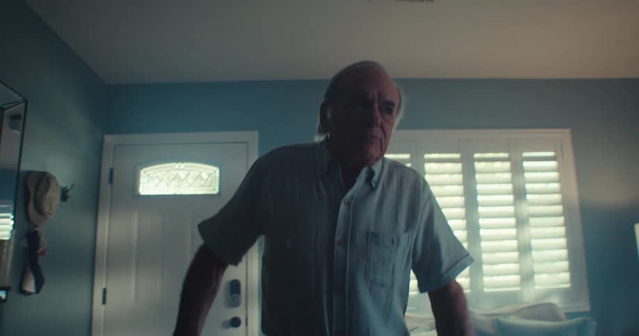 An elderly gentleman dances around in his living room as sunlight filters through the window blinds in the background
